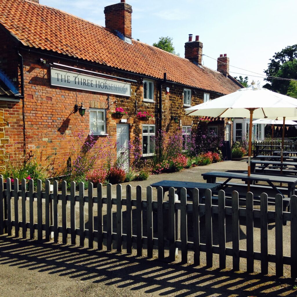 "Exterior of The Three Horseshoes pub with red brick walls, tiled roof, and a cosy outdoor seating area featuring picnic benches and white umbrellas. Vibrant flowers decorate the front, bordered by a wooden picket fence on a sunny day."